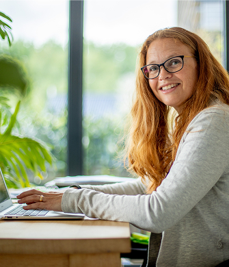Een vrouw met rood haar en een vril zit achter de compute en kijkt zijwaarts en in de camera.