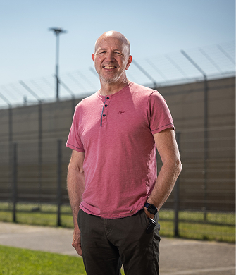 Man staat buiten, draagt een rood t-shirt, hij lacht en kijkt in de camera. Het is zonnig.