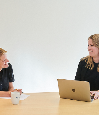 Twee vrouwen met elkaar in gesprek. Een vrouw staat en de ander zit. Ze kijken elkaar aan.
