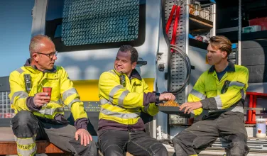 Three workers in yellow safety jackets sitting outside near a work van, taking a break and sharing a snack.