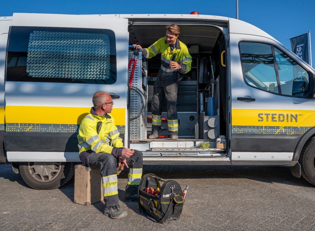 Two Stedin utility workers wearing safety helmets and high-visibility jackets inspect electrical panels inside a facility.
