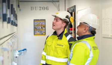 Two Stedin utility workers wearing safety helmets and high-visibility jackets inspect electrical panels inside a facility.