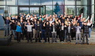 Große Gruppe von Auszubildenden steht vor dem Hager Unternehmensgebäude in Blieskastel und winkt in die Kamera.