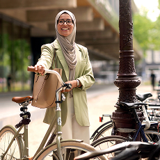 Lachende vrouw met hijab en bril staand bij een fiets op een stadsstraat.