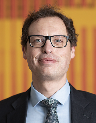 Een man met bril en pak, glimlachend voor gele achtergrond.