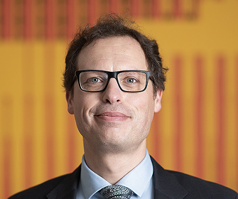 Een man met bril en pak, glimlachend voor gele achtergrond.