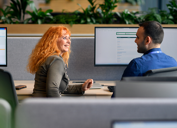 Two colleagues discussing work at a desk with multiple computer monitors in an office setting.