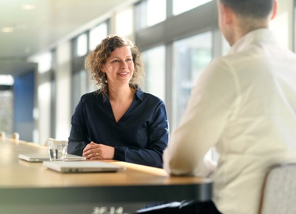 A professional woman smiling while conversing with a colleague across a table in a bright office.