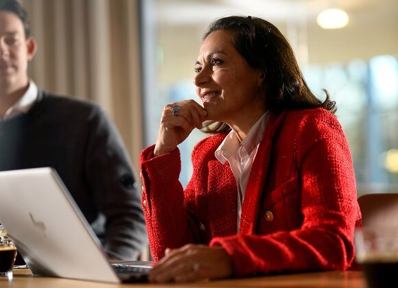 A smiling woman in a red blazer sitting at a desk with a laptop during an office meeting.