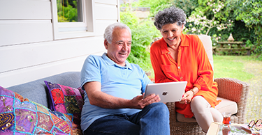 Twee personen zitten buiten op een terrasbank en bekijken samen een tablet, met een tuin op de achtergrond.