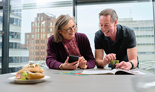 Twee personen zitten aan een tafel met een smartphone en documenten tijdens een informeel overleg, met lunch op de voorgrond.