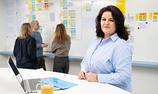 Persoon staat bij een laptop voor een wand vol scrumborden waar anderen overleggen.