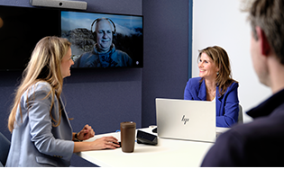 Personen zitten in een vergaderruimte met een videogesprek op het scherm.