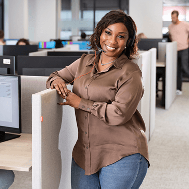 Persoon staat bij een werkplek in een kantoorruimte waar anderen aan het werk zijn.