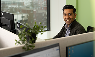 Persoon zit aan een bureau in een kantoorruimte met meerdere computerschermen en een groot raam met uitzicht op de stad.