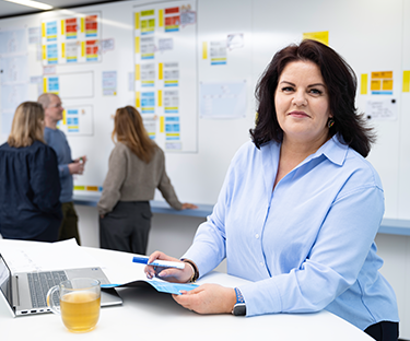 Persoon staat bij een laptop voor een wand vol scrumborden waar anderen overleggen.