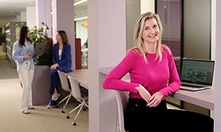 Persoon zit aan een werkplek met een open laptop, terwijl twee anderen met elkaar praten in de achtergrond van een moderne kantoorruimte.