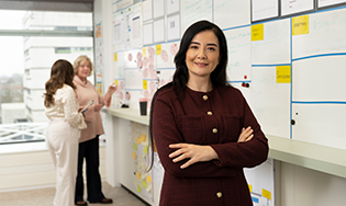 Persoon staat met armen gekruist voor een dagstartbord, terwijl twee anderen samenwerken aan de achtergrond.
