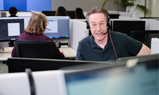 Persoon werkt met een headset aan een bureau in een kantoor met meerdere computerschermen.