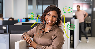 Persoon staat bij een werkplek in een kantoorruimte waar anderen aan het werk zijn.