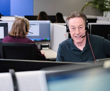 Persoon werkt met een headset aan een bureau in een kantoor met meerdere computerschermen.