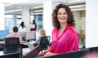 Persoon werkt aan een bureau in een open kantoorruimte met collega’s op de achtergrond.