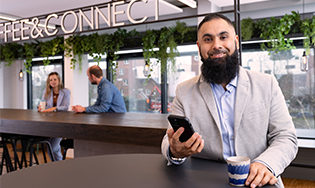 Persoon zit aan een tafel met een smartphone en koffiekopje, in een ruimte met een Coffee & Connect‑bord en anderen op de achtergrond.