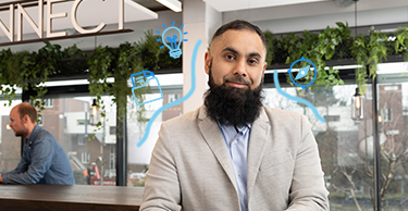 Persoon zit aan een tafel met een smartphone en koffiekopje, in een ruimte met een Coffee & Connect‑bord en anderen op de achtergrond.