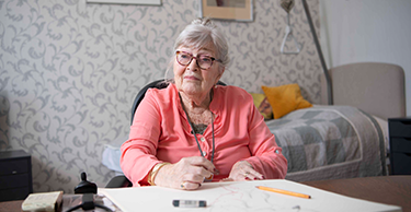 Persoon zit aan een tafel in een kamer met een bed op de achtergrond en werkt aan een activiteit op tafel.