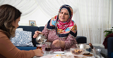 Persoon schenkt thee in voor iemand anders in een woonkamer met een bank en kussens.