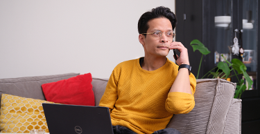 Persoon zit op een bank met een laptop en telefoneert in een woonkamer.