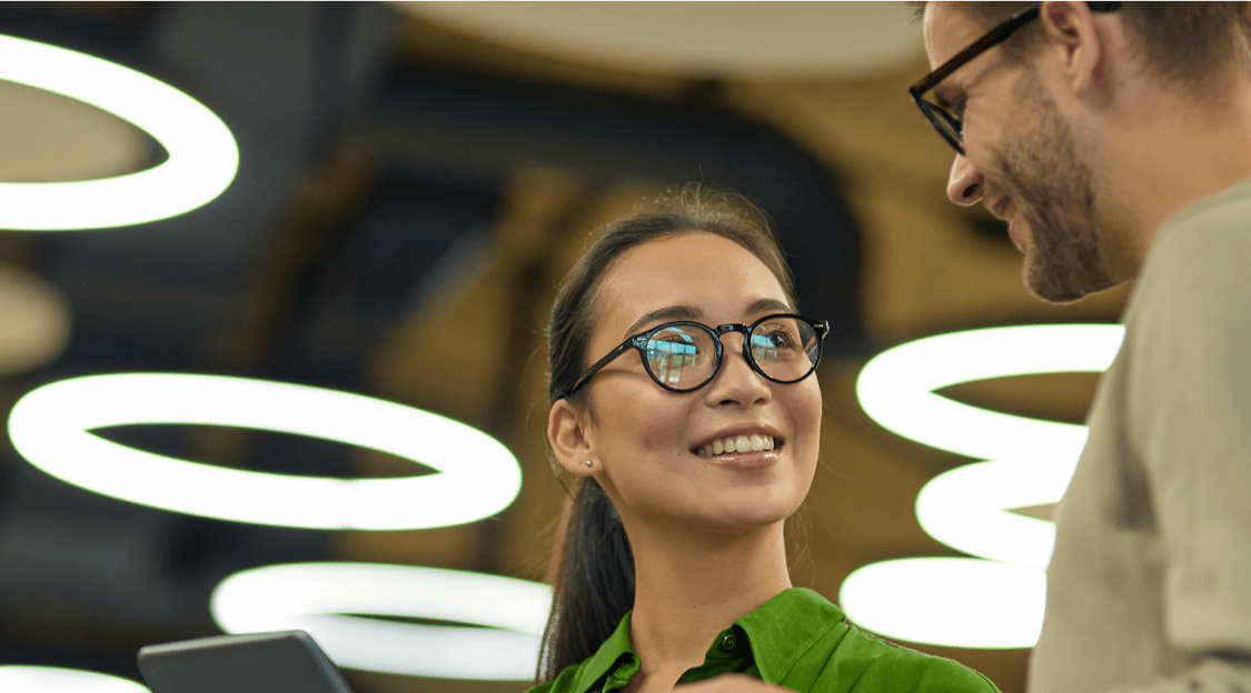 Woman in glasses and green shirt holding tablet while talking to another colleague