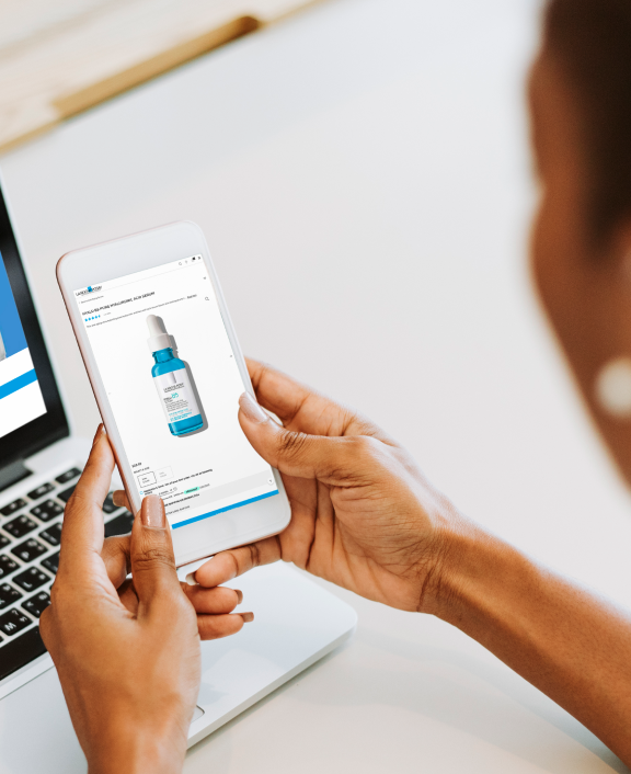 Person holding smartphone browsing skincare product page beside laptop