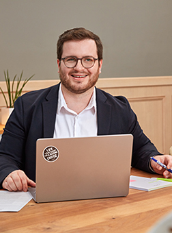 Personne portant des lunettes, utilisant un ordinateur portable à son bureau et souriant dans son espace de travail.