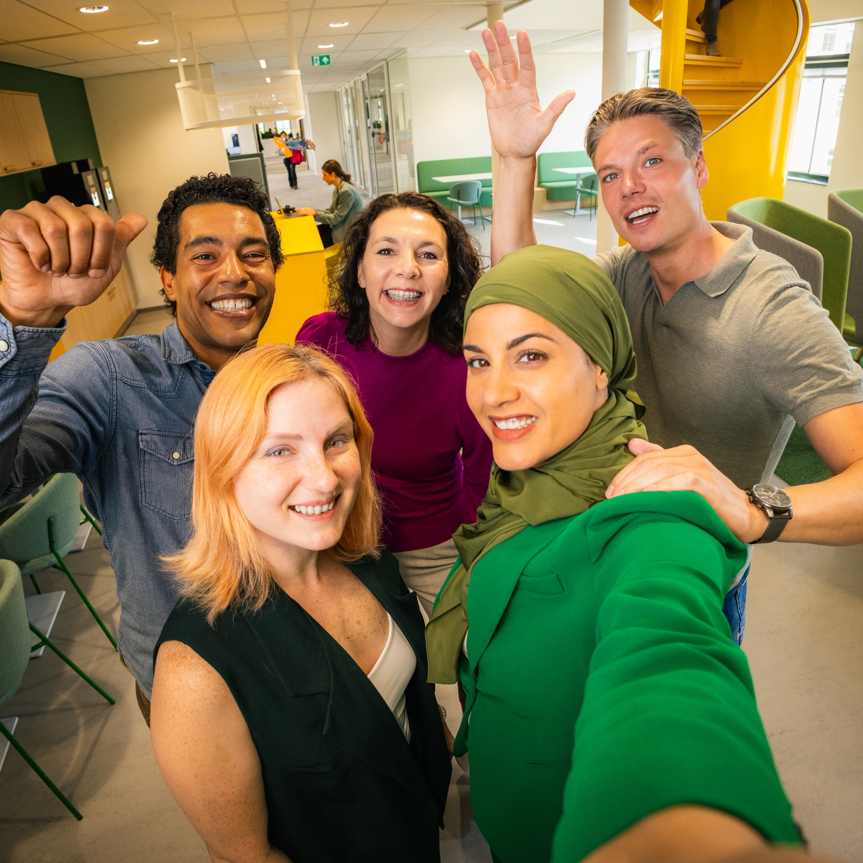 Diverse groep collega’s maakt een groepsselfie in een moderne kantooromgeving, met werkplekken, zitgedeeltes en een opvallende gele trap op de achtergrond.