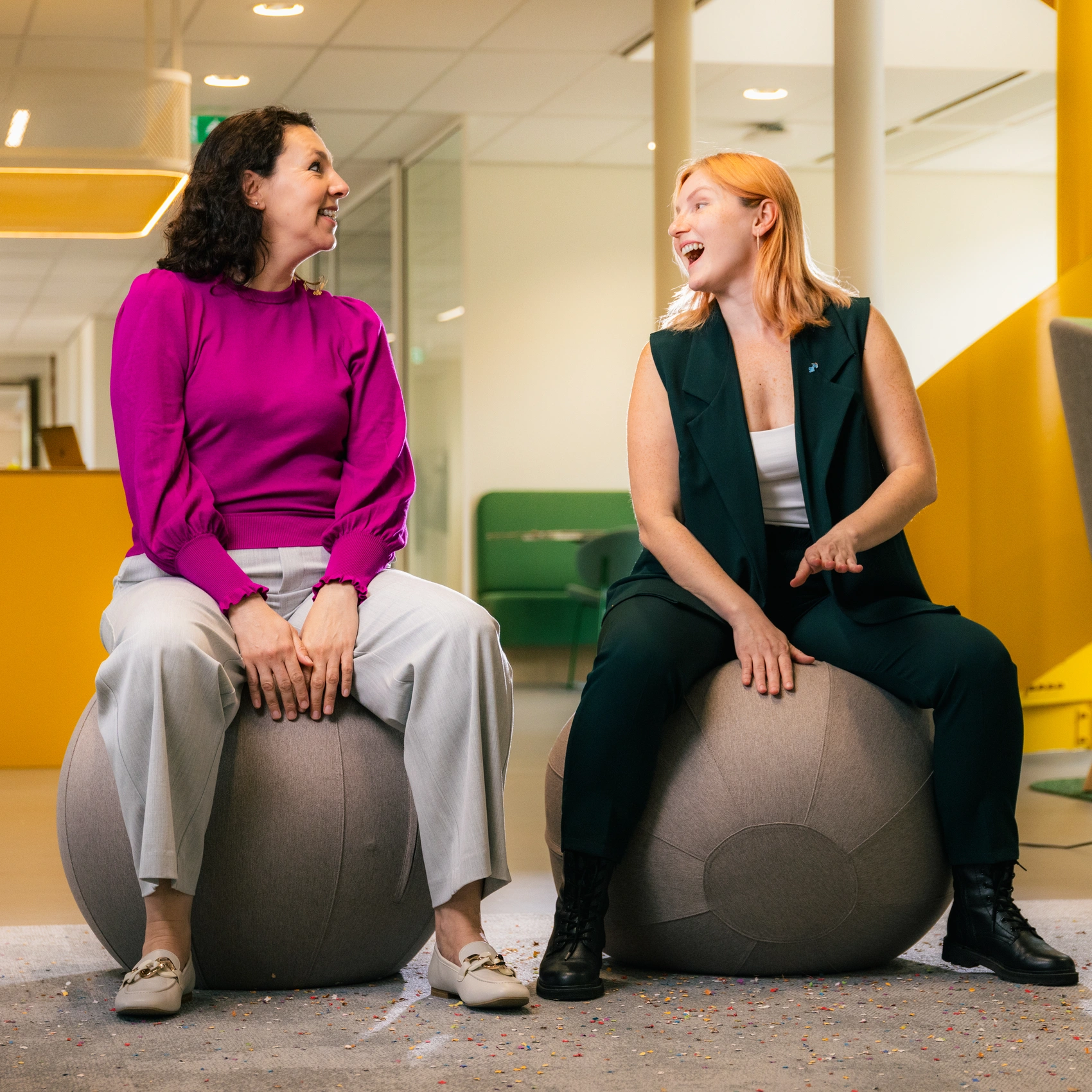 Twee collega's zitten op grote zitballen in een moderne kantooromgeving en voeren een gesprek, met werk- en ontmoetingsruimtes op de achtergrond.