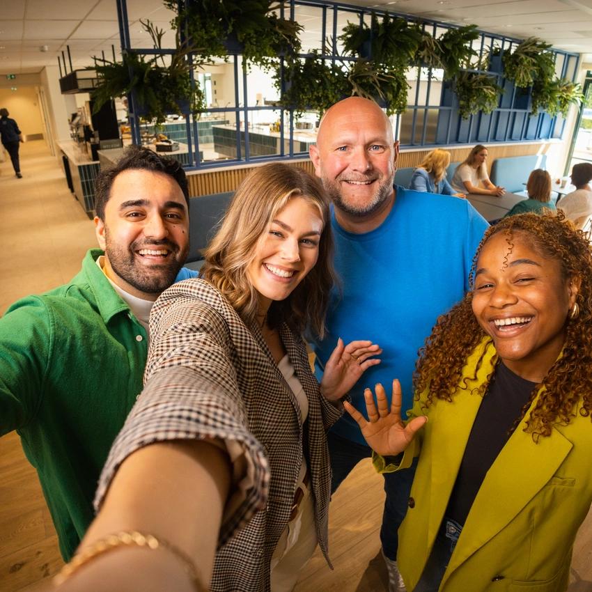 Vier collega’s lachen en maken een selfie in een moderne kantoorlounge.