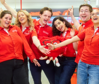Groep winkelmedewerkers in rode bedrijfskleding poseert samen in een drogisterijwinkel tussen de winkelrekken.
