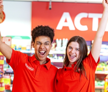 Twee winkelmedewerkers in rode bedrijfskleding vormen een hart met hun handen in een drogisterijwinkel.